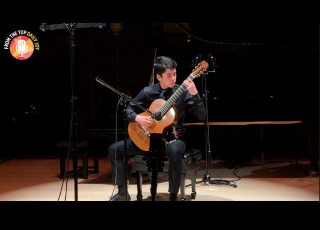 boy sits on stage playing guitar in front of a microphone.