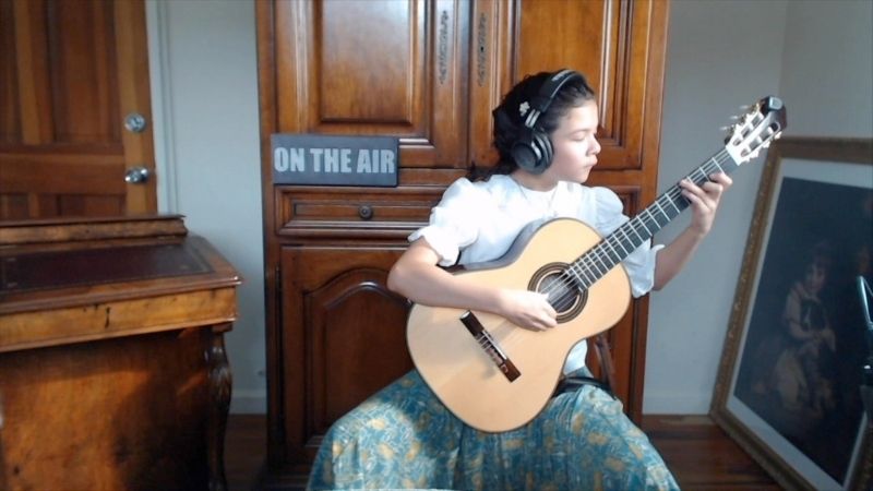 young girl plays guitar in a house in front of a sign that says 'on the air'.