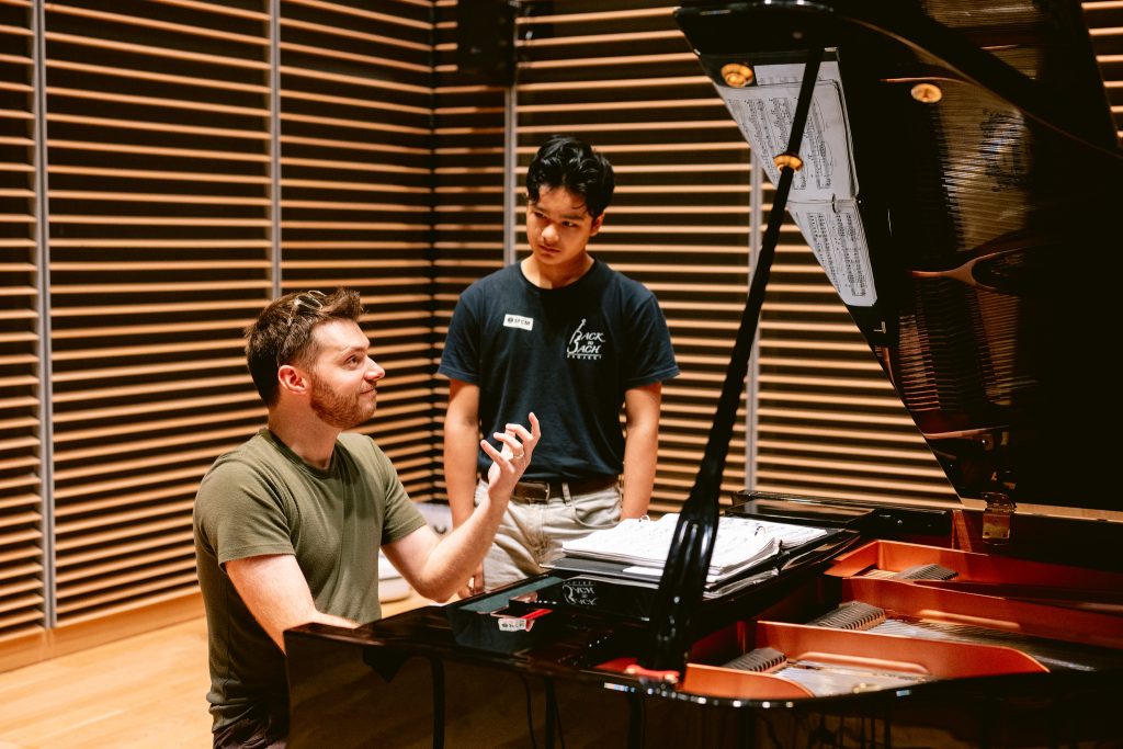 Man sits at piano and gestures with hand while teen boy looks on